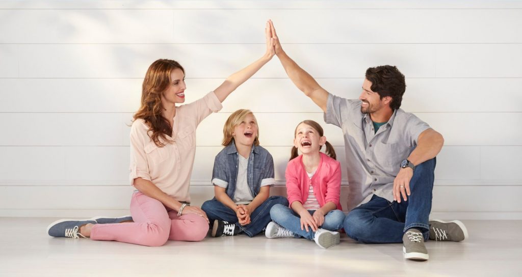 Husband and wife smiling with their son and daughter. Parents arms reaching above children and joined together creating the shape of the roof of a house.