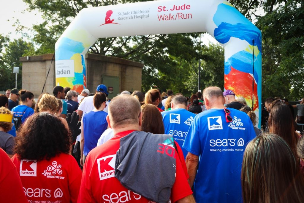 Members of the Kmart and Sears St. Jude Walk/Run team join a crowd of walkers crossing the start line to the 2019 walk run event wearing red and blue T-shirts.