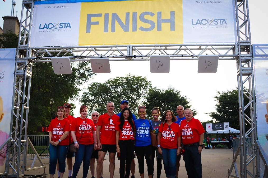 Seven Sears and Kmart associates stand in a line smiling under the St. Jude Walk/Run finish line.