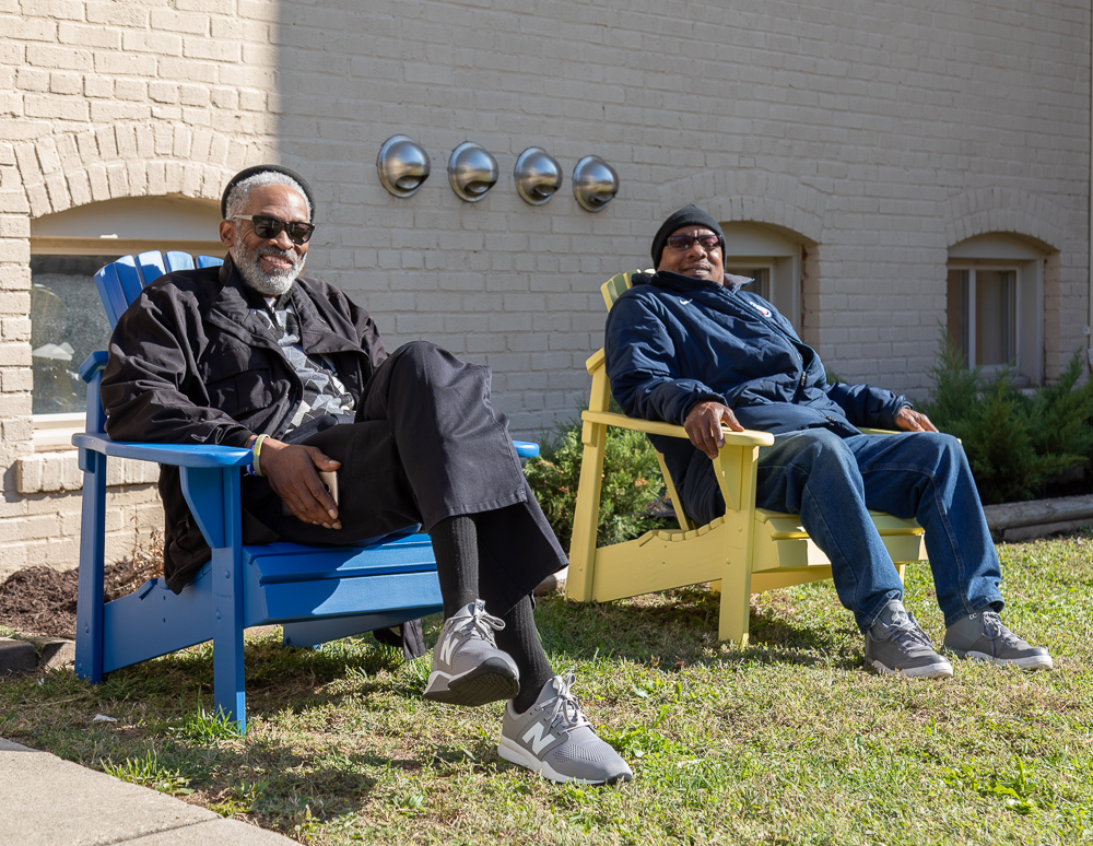 Two men smile as they lounge in bright blue and yellow Adirondack chairs on a grassy lawn of a tan brick building.