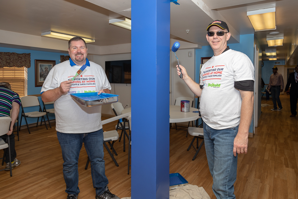 Two volunteers smile for the camera after painting a blue support beam. One is holding a brush and paint pan of blue paint matching the beam while the other is painting with a roller. Both are wearing Sears Heroes at Home and Rebuilding Together T-shirts. 