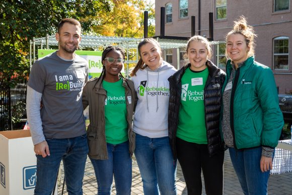 Five volunteers wearing various green and white Rebuilding Together apparel smile in front of a sunny Rebuilding Together and Heroes at Home project site that was supported in part by Sears