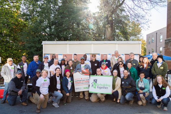 A large group of people stand with signage for Rebuilding Together and Heroes at Home in front of a project site.